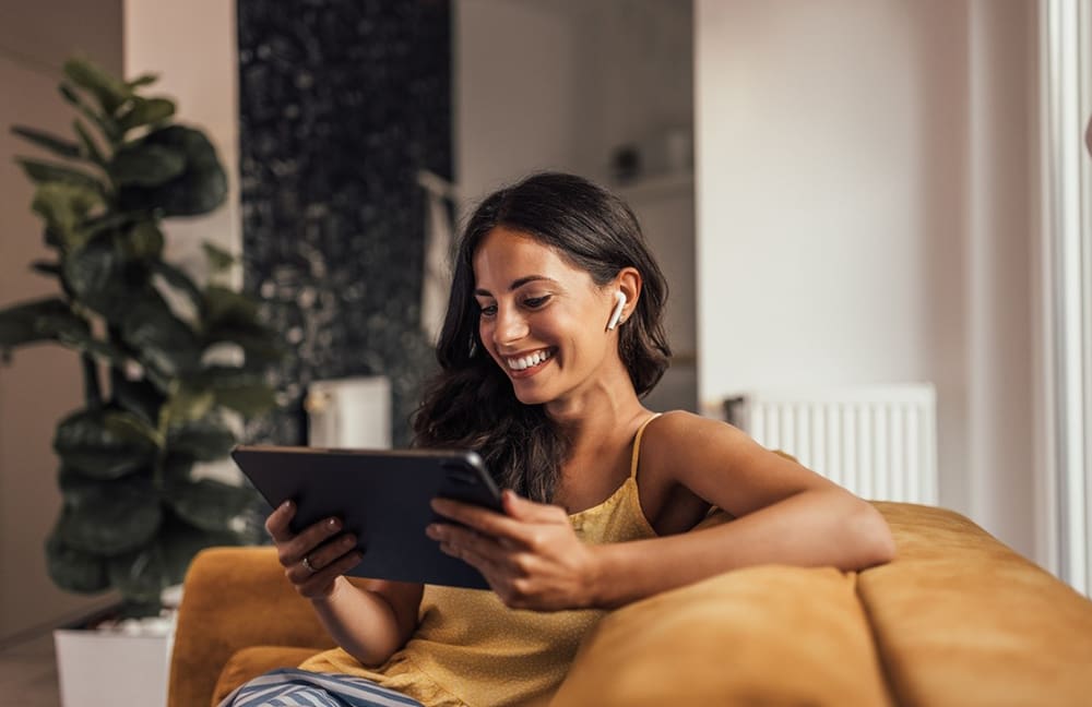 Eine Frau sitzt lachend mit einem Tablet in der Hand auf dem Sofa.