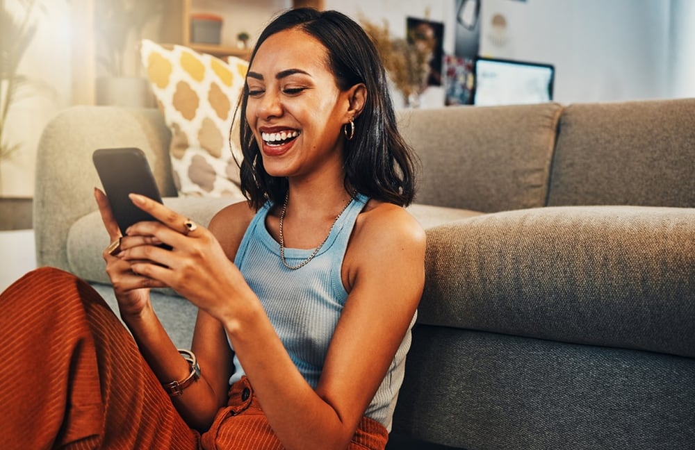 Femme souriante assise devant un canapé, un téléphone mobile à la main.