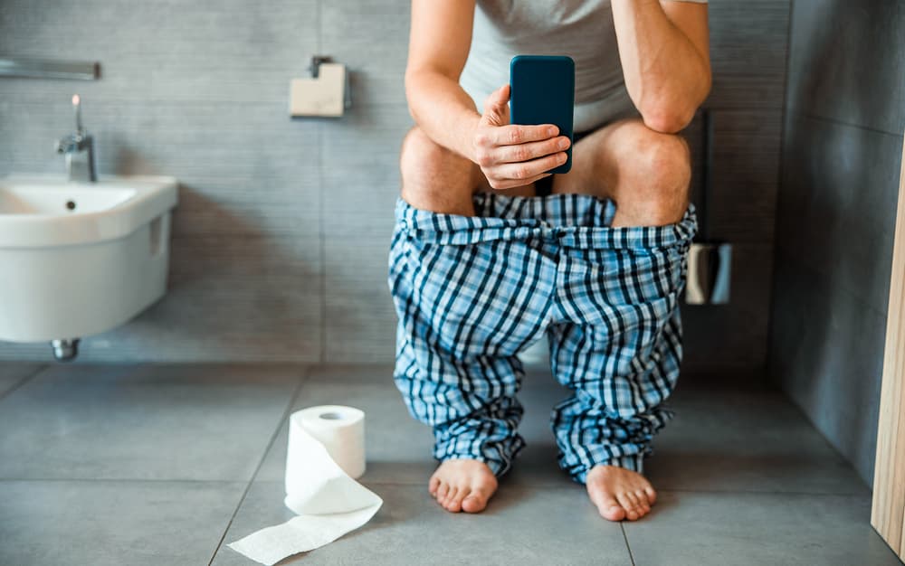 Un mec est assis sur les toilettes avec son téléphone mobile à la main.