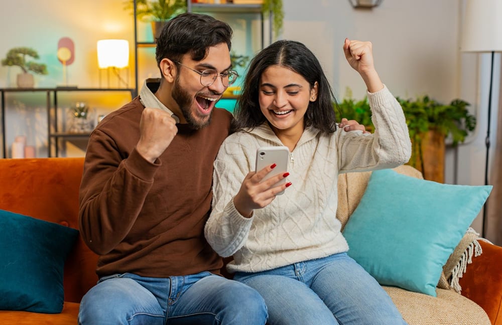 Ein Mann und eine Frau mit Handy in der Hand jubeln sitzend auf einem Sofa