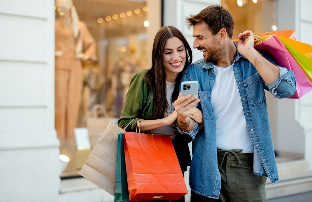 Un homme avec un téléphone mobile à la main et une femme avec des sacs de courses devant un magasin
